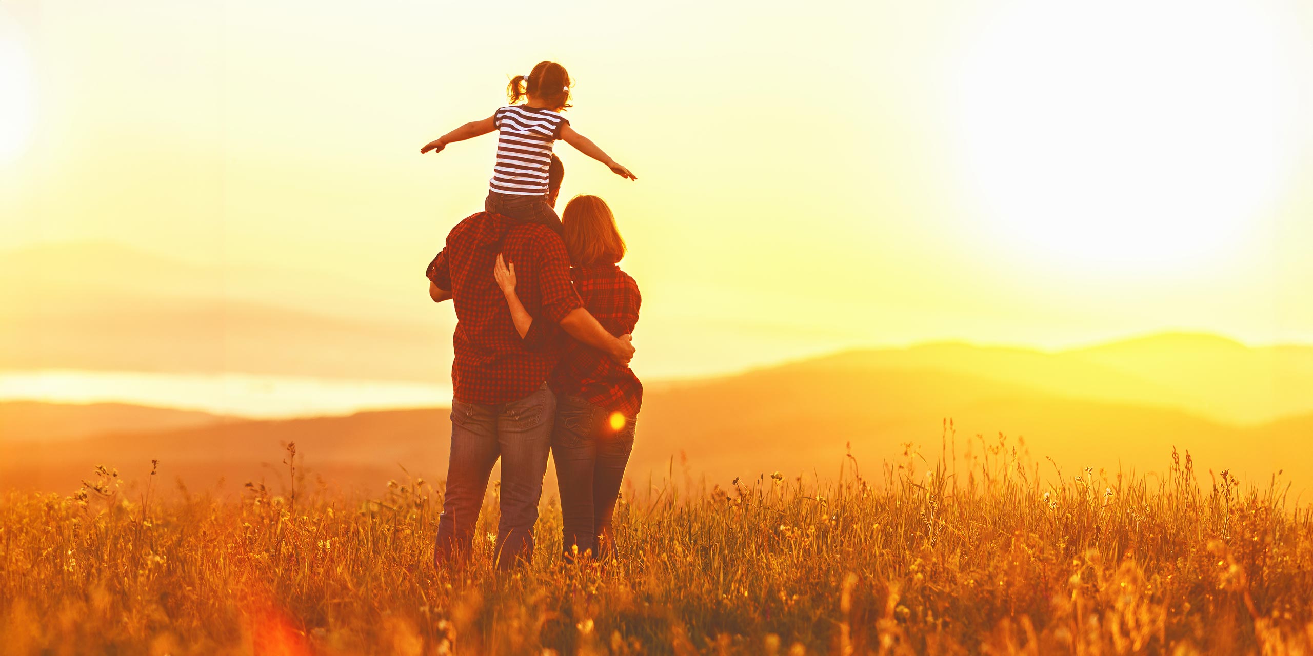 Eine junge Familie im Sonnenuntergang, den Rücken zur Kamera. Die Tochter sitzt auf den Schultern des Vaters.